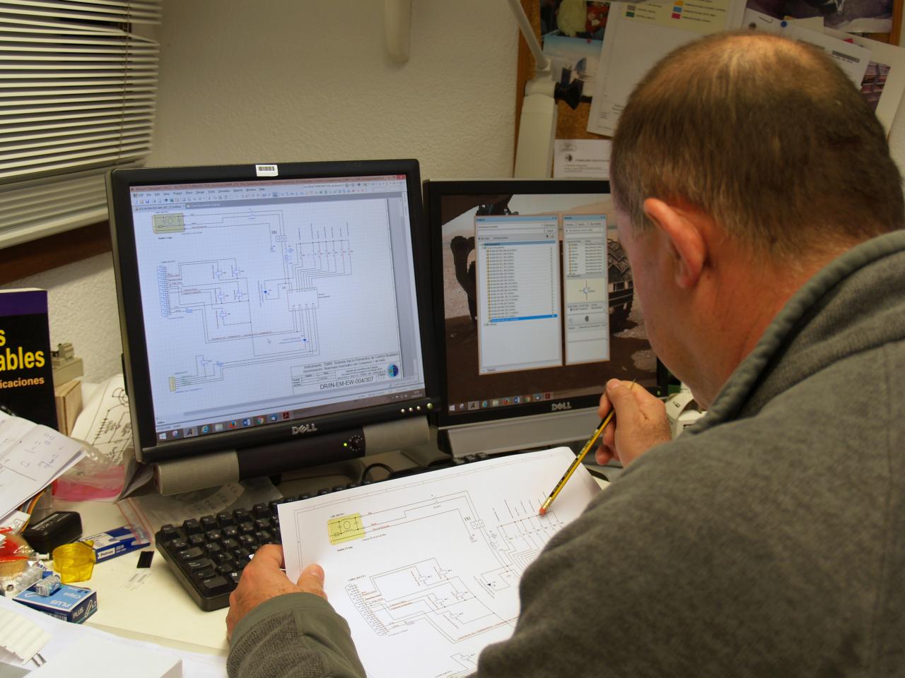 Vista de un técnico trabajando en un diseño electrónico con un plano y la pantalla del ordenador