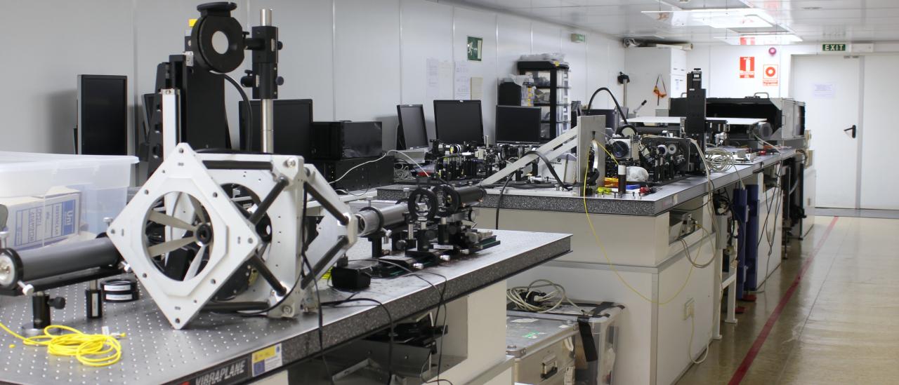 Vista general de una de las salas (A) del laboratorio de óptica. Laboratorio alargado con mesas metálicas con agujeros y diversos elementos ópticos y mecánicos encima de ellas