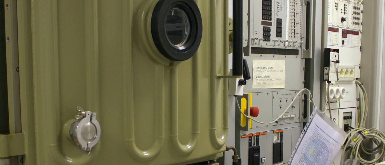 Vista de la evaporadora y de su electrónica asociada en el laboratorio. Máquina con una puerta reforzada y un ojo de buey, y dos grandes racks con componentes electrónicos, botones e indicadores para el control de la máquina.