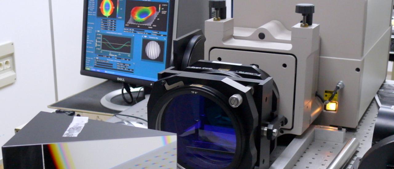 Vista del interferómetro en una mesa del laboratorio. Dipositivo optoelectrónico con una gran prisma y una lente delante y una pantalla de ordenador con gráficos detrás