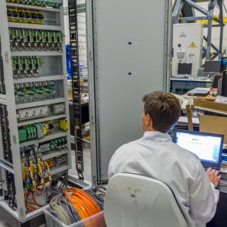 Imagen de un ingeniero trabajando en la electrónica del sistema de óptica adaptativa para GTC en el laboratorio. Ingeniero sentado trabajando en un ordenador con un armario de electrónica abierto a su lado