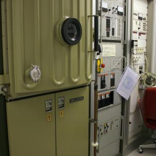 Vista de la evaporadora y de su electrónica asociada en el laboratorio. Máquina con una puerta reforzada y un ojo de buey, y dos racks de gran tamaño con componentes electrónicos, botones e indicadores para el control de la máquina