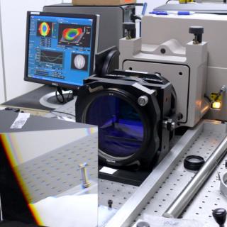 Vista del interferómetro en una mesa del laboratorio. Dipositivo optoelectrónico con una gran prisma y una lente delante y una pantalla de ordenador con gráficos detrás